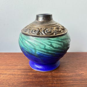 Puede incluir: Un pequeño tarro de cerámica decorativo con un patrón arremolinado de esmalte azul y verde. El tarro tiene una tapa de metal plateado con un diseño ornamental en relieve. La base es de color azul sólido.
