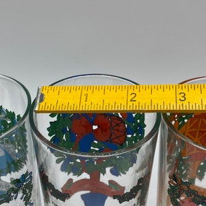 May include: Three clear drinking glasses with colorful holiday-themed designs. Each glass features a different illustration, including a drum, a tree, and a geometric pattern. The glasses are approximately 3 inches in height.