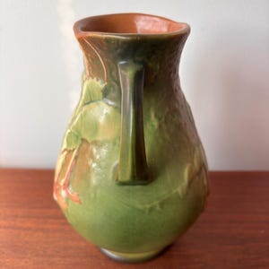 May include: A green ceramic jug with a handle, decorated with a leaf pattern. The interior is brown, and the surface is textured. The handle is a darker shade of green.