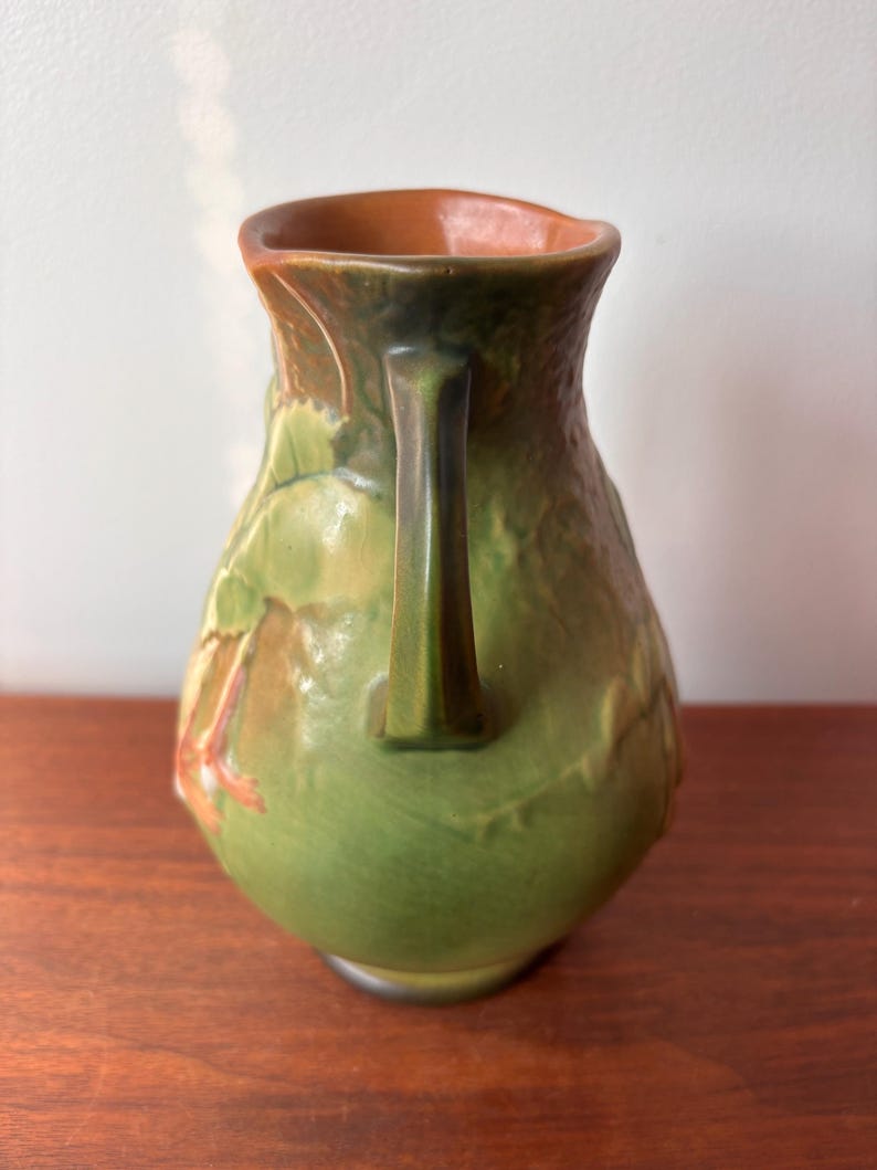 May include: A green ceramic jug with a handle, decorated with a leaf pattern. The interior is brown, and the surface is textured. The handle is a darker shade of green.