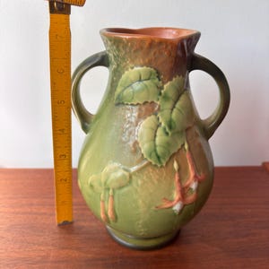 May include: A green ceramic vase with two handles and a textured surface. The vase features embossed leaves and flowers in shades of green, orange, and white. The vase is approximately 18 cm tall.