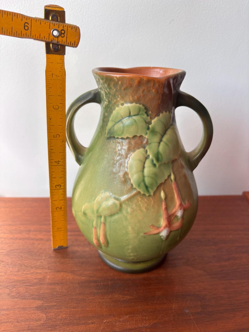 May include: A green ceramic vase with two handles and a textured surface. The vase features embossed leaves and flowers in shades of green, orange, and white. The vase is approximately 18 cm tall.