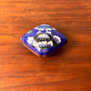 Puede incluir: Una caja decorativa azul en forma de diamante con un diseño floral. La caja presenta un fondo azul oscuro con flores blancas y negras, delineadas en oro. Los bordes también están adornados con oro.