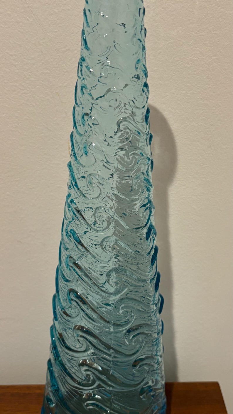 May include: A tall, clear blue glass vase with a swirling, textured design. The vase is shaped like a cone and has a wide base.