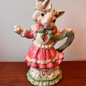 May include: A ceramic teapot featuring two bunny figures, one wearing a top hat and the other a floral headpiece. The teapot is primarily pink and white, with green and gold accents. The bunnies are embracing, and the teapot has a decorative base.