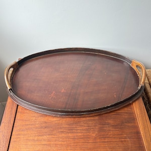 May include: An oval-shaped wooden serving tray with a dark brown finish and a smooth, flat base. The tray features two integrated curved handles on opposite sides, showing visible wood grain and minor surface wear consistent with age and use.