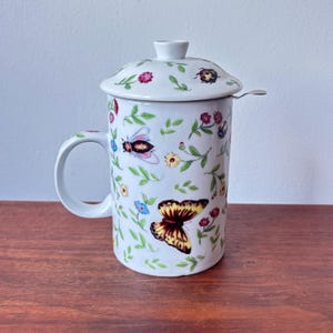 Può includere: Tazza da tè in ceramica bianca con coperchio e infusore, ornata da un colorato motivo floreale e di insetti. Il design include farfalle, api e fiori nei toni del giallo, rosso e blu. La tazza ha un manico e un infusore in acciaio inossidabile.