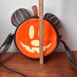 May include: Orange pumpkin-shaped crossbody bag with a smiling jack-o'-lantern face. The bag has black Mickey Mouse ears with orange stripes and a brown stem. The bag is approximately 8 inches tall.