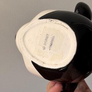 May include: A black and white ceramic mug with a cartoon character design. The mug has a glossy finish and a rounded shape. The bottom of the mug has a sticker with the word "Disney" and a copyright symbol.