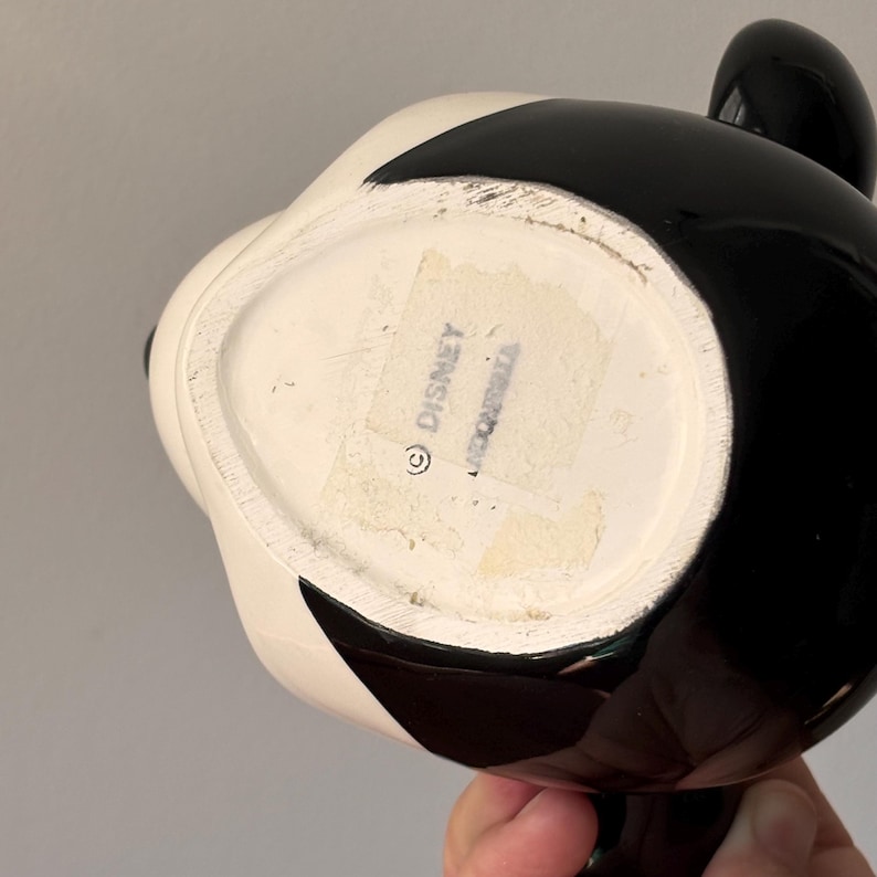 May include: A black and white ceramic mug with a cartoon character design. The mug has a glossy finish and a rounded shape. The bottom of the mug has a sticker with the word "Disney" and a copyright symbol.