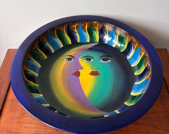 May include: A hand-painted ceramic bowl with a celestial design. The bowl features a crescent moon and sun face in shades of yellow, purple, and green, with red lips and blue eyes. The rim is a deep blue with a decorative border of blue, green, and yellow.