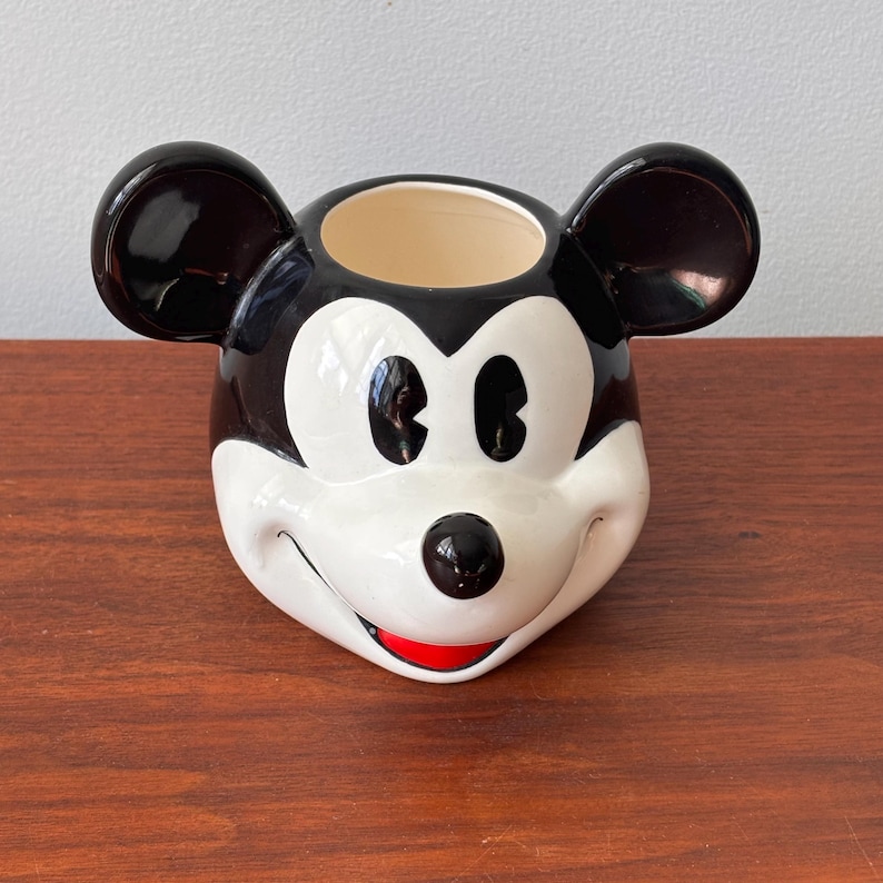 May include: A ceramic Mickey Mouse head-shaped container. The container features black ears, a black and white face, and a red mouth. The interior is beige. The container is sitting on a brown surface.