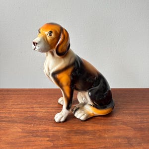 Peut inclure: Une figurine en céramique représentant un chien de chasse assis avec une finition brillante. Le chien présente un pelage tricolore avec des taches noires, fauves et blanches. Il a de longues oreilles tombantes, un museau blanc et un regard concentré.
