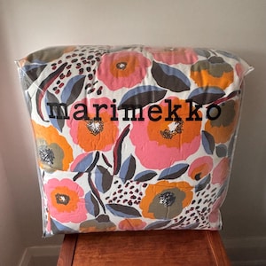 Op de afbeelding: Een wit en roze kussen met een bloemenpatroon en de tekst "marimekko" in zwarte letters.