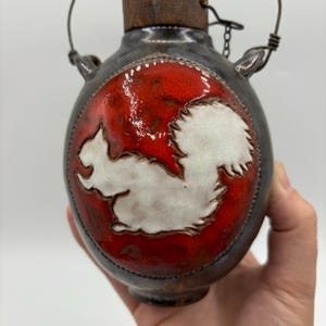 May include: A small ceramic bottle with a red and white squirrel design. The bottle has a cork stopper and a wire handle.