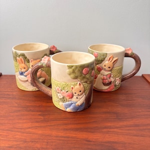 Puede incluir: Tres tazas de cerámica con diseños en relieve de conejos y ositos de peluche en un jardín. Las tazas tienen cuerpos color crema, asas marrones con detalles de flores rosas e ilustraciones detalladas. Las tazas están sobre una superficie de madera.