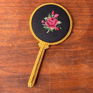 Puede incluir: Un espejo de mano con marco dorado y fondo de tela negra con una rosa rosa bordada. El espejo tiene un mango dorado con un diseño de cuerda.