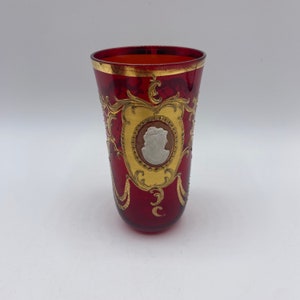 Puede incluir: Un vaso rojo de vidrio con detalles pintados en oro y blanco. El vaso presenta un camafeo de un perfil de mujer en un marco dorado.