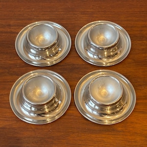 May include: Four silver egg cups with a classic design. Each cup has a rounded bowl shape set on a circular base. The cups are made of a shiny, reflective metal and are arranged on a wooden surface.
