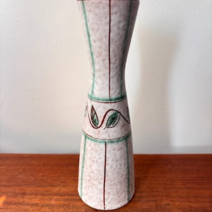 May include: A tall, decorative vase with a speckled, off-white finish. It features vertical green stripes, a central red stripe, and a band of leaf designs. The vase has a flared top and a narrower midsection, standing on a wooden surface.