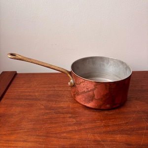 Puede incluir: Una pequeña cacerola de cobre con un asa de latón. El interior de la cacerola es de color plateado. El exterior de cobre tiene una pátina marrón rojiza. El asa está sujeta a la cacerola con un remache de latón. La cacerola está sobre una superficie de madera.