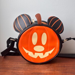 May include: A round, orange pumpkin-shaped crossbody bag with a black trim and strap. The bag features a smiling jack-o'-lantern face and black Mickey Mouse ears with orange stripes. A brown stem is at the top, and a gold zipper is visible.
