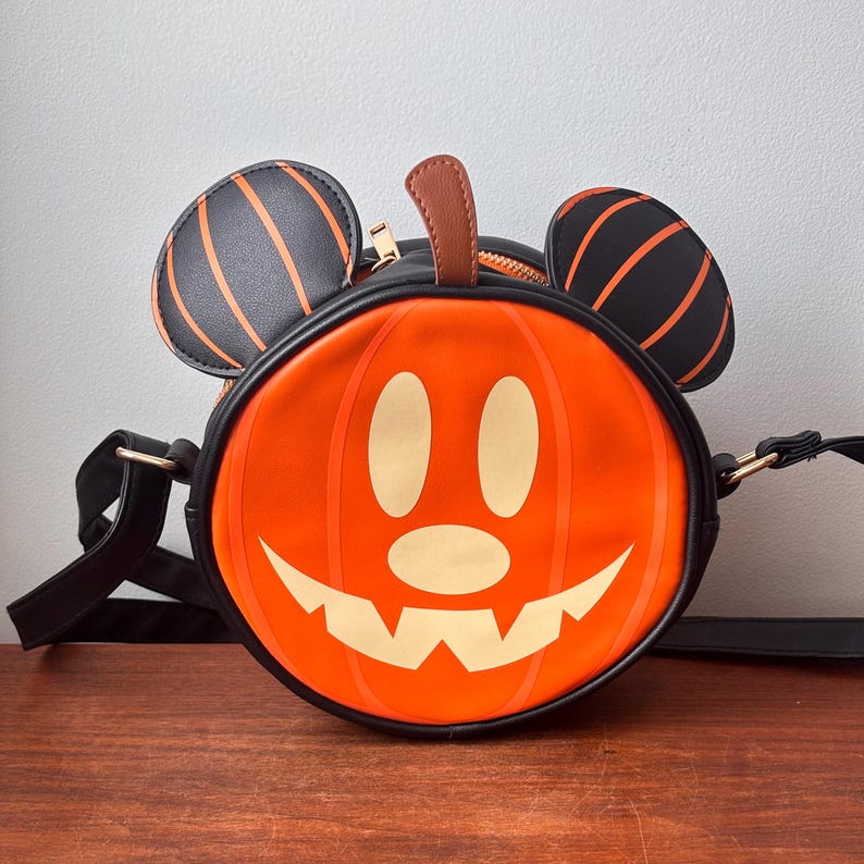 May include: A round, orange pumpkin-shaped crossbody bag with a black trim and strap. The bag features a smiling jack-o'-lantern face and black Mickey Mouse ears with orange stripes. A brown stem is at the top, and a gold zipper is visible.