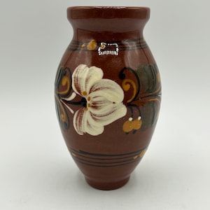 Puede incluir: Un pequeño jarrón de cerámica marrón con una flor blanca y hojas verdes pintadas en el lateral. El jarrón tiene un esmalte marrón y una forma ligeramente redondeada.