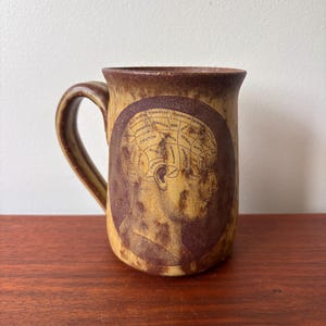 Puede incluir: Una taza de cerámica con un acabado moteado marrón y beige. La taza presenta una ilustración circular de una cabeza humana con un diagrama del cerebro. El asa es marrón y la taza está sobre una superficie de madera.