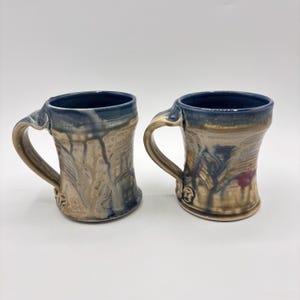 May include: Two handmade ceramic mugs with blue and brown glaze. The mugs have a unique, textured design and a handle.