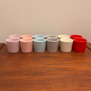 Op de afbeelding: Een verzameling van twaalf kleine, cilindervormige keramische potten in verschillende pastelkleuren, waaronder roze, perzik, blauw, grijs, crème en rood. De potten zijn in twee rijen op een houten oppervlak geplaatst, wat hun eenvoudige, moderne ontwerp laat zien.