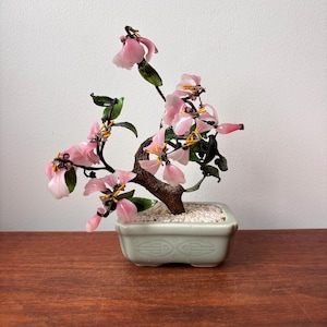 May include: A miniature bonsai tree with pink blossoms and green leaves. The tree is planted in a pale green rectangular pot filled with white pebbles. The bonsai's trunk is brown, and the flowers are a delicate pink.