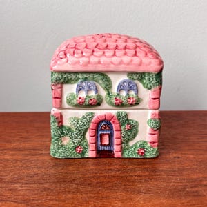 Peut inclure: Un récipient en céramique en forme de maison avec un toit rose et des murs blancs. La maison a des détails de fenêtres bleues, une porte rouge et est décorée de vignes vertes et de fleurs rouges. Le récipient est posé sur une surface en bois.