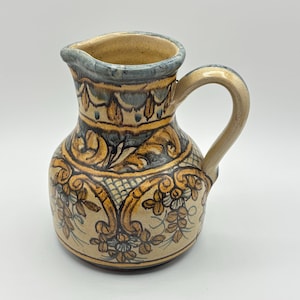 Könnte beinhalten: Ein kleiner, handbemalter Keramik Krug mit einem blauen, gelben und braunen Blumendesign. Der Krug hat einen runden Körper und einen Henkel.