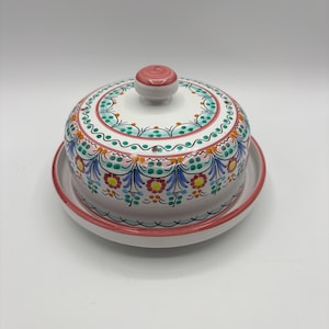 May include: A white ceramic cheese dome with a pink lid and a colorful floral design. The dome has a matching base.