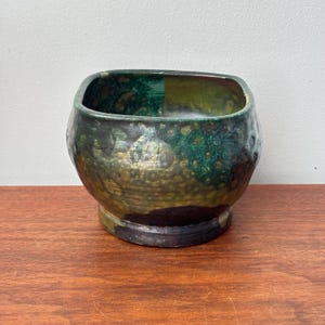 May include: A rounded ceramic bowl with a wide opening and a thick base. The exterior features a mottled glaze with blended shades of deep forest green, earthy brown, and metallic gold, creating a textured appearance across the curved surface.