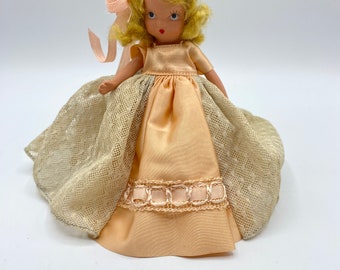 Puede incluir: Una muñeca de porcelana vintage que lleva un vestido de satén melocotón con una superposición de encaje y una cinta rosa. La muñeca tiene el pelo rubio y los ojos azules.