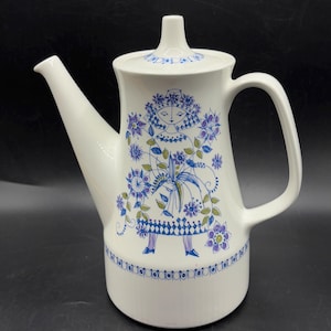 May include: White ceramic teapot with a decorative floral and figure design in blue, purple, and green. The teapot features a long spout, a curved handle, and a lid with a small knob. The design includes a stylized figure and floral patterns.