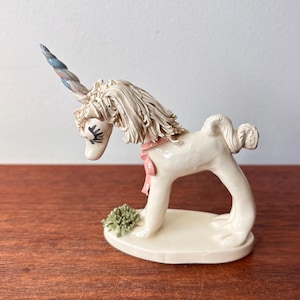 Op de afbeelding: Een crèmekleurig keramieken eenhoornbeeldje met een roze lint en een veelkleurige hoorn. De eenhoorn heeft een vloeiende manen en een klein groen struikje aan de basis. Het beeldje staat op een witte ovale basis.