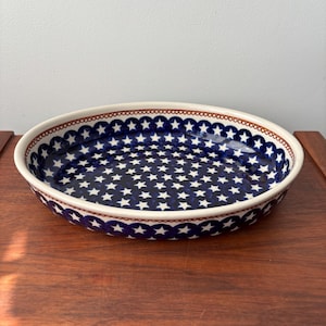 Pode incluir: Um grande prato de cozedura oval de cerâmica com um padrão de estrelas brancas sobre um fundo azul escuro. O prato tem uma borda castanha com uma borda festoneada.
