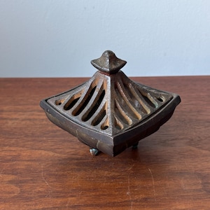 May include: A square, dark-toned metal incense burner featuring a removable lid with a slatted, geometric design and a pointed finial. The base is supported by small, short feet, and the overall surface displays a weathered, textured finish.
