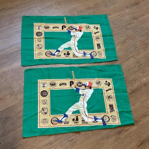 May include: Two green pillowcases with a baseball player in a white uniform with a red and blue hat. The player is holding a bat and is surrounded by a frame of baseball team logos. The pillowcases are made of a soft, comfortable fabric.