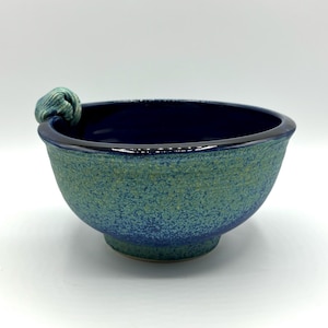May include: A blue and green ceramic bowl with a textured finish. The bowl has a unique handle that is shaped like a knot.