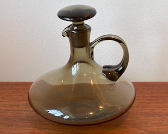 May include: A smoke-colored glass decanter with a wide, rounded base, a slender neck, and a curved handle. The vessel features a matching circular glass stopper that fits securely into the top opening, creating a cohesive and functional design.
