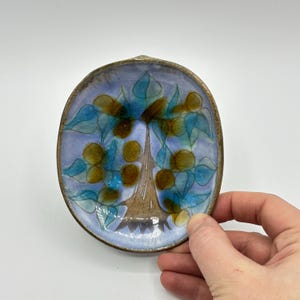 May include: A small, oval ceramic dish with a blue glaze and a hand-painted design of a tree with brown branches and blue and yellow leaves.