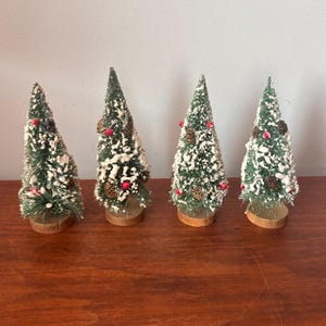 May include: Four miniature Christmas trees with green bottle brush needles, white snow, red berries, and small pine cones. The trees are mounted on wooden bases, creating a festive holiday decoration.
