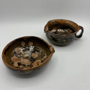 May include: Two handmade ceramic bowls with a brown and tan speckled glaze. One bowl has a handle and a spout, while the other is a simple bowl with a spout. The pottery has a rustic, earthy aesthetic.