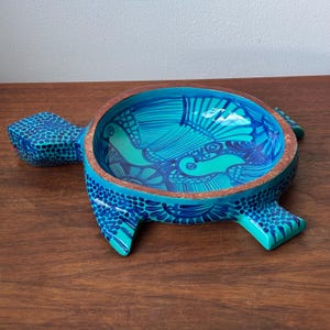 May include: A turtle-shaped bowl featuring a vibrant turquoise and dark blue hand-painted pattern on its exterior and interior. The bowl has a natural wood-toned rim and a detailed shell design with intricate line work and dot motifs.