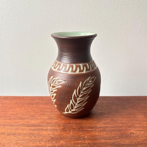 May include: A small brown ceramic vase with a light green glaze on the rim. The vase has a carved design of leaves and a geometric pattern around the middle.