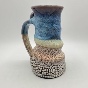 Puede incluir: Una taza de cerámica hecha a mano con un esmalte azul, blanco y marrón. La taza tiene un diseño único y texturizado y una amplia asa redondeada.
